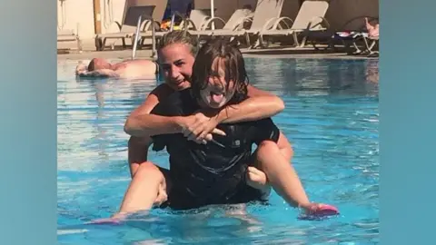 Family photo Carolyn Cox is pictured being carried by Ellis on his back in a swimming pool. She is smiling at the camera and Ellis is smiling and sticking his tongue out.