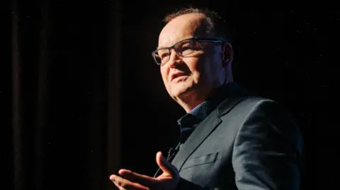 Tony Moretta pictured on stage giving a talk in a darkened room. He is wearing a dark suit and glasses. 