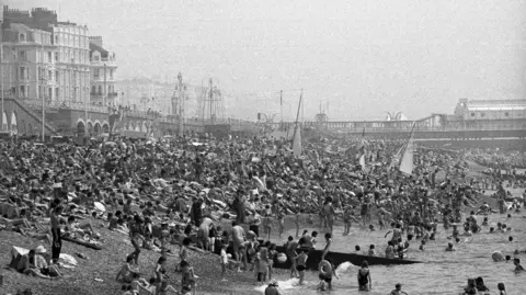 PA Media Hundreds of people covering Brighton beach and in the water. The pier and shoreline are visible in the black and white image.