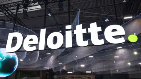 A large Deloitte sign in white letters with a small green full stop at the end. The large lighted sign is hanging over a large hall floor and there is a pixelated globe on the left of the picture.