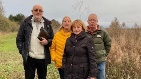 Four people, two men and two women, are standing side by side on the common, they are wearing coats and the sky is grey.