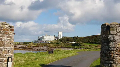MANX SCENES The exterior of the hotel from when it was in use, the land is well-kept, there are cars parked, and you can see the white building with a tower in the background.