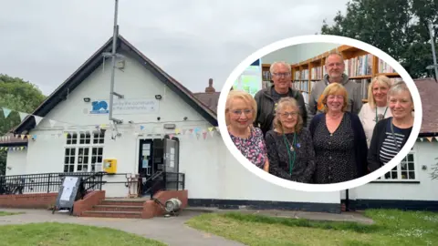 A white building with multicoloured bunting strung up in front. The building has a sign that reads 'Anlaby Park Library' and 'Run by the community for the community'. The library has a black front door which is open, and has red steps and a disabled ramp leading up to it. Above, the sky is cloudy and grey and you can see green trees in the background. Below is a path and green grass with white flowers on. To the right is a white oval featuring a group picture of five women and two men in a library setting, with a long brown bookshelf behind them filled with books.