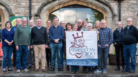 A group of men and women who are the Athelstan 1100 Big Dig project team holding a banner advertising their events this summer