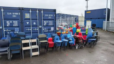 Hull City Council Row of stacked chairs of various types and colours stand in front of two dark blue shipping containers 