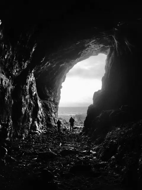 Bill Ritchie A black-and-white view from inside a large cave, showing two people silhouetted against the bright light outside.
