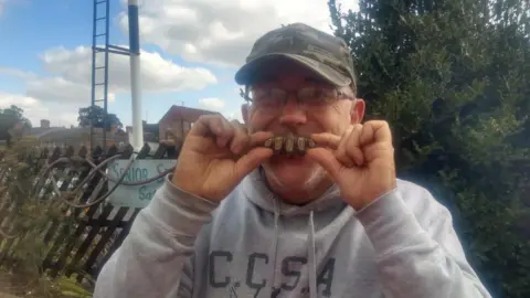 Andy Green Andy Green holding up the sword pommel to his mouth, where its five lobes look a bit like five large teeth. He is smiling and wearing a baseball hat, glasses and a grey sweatshirt. Behind him is a fence, trees and houses.