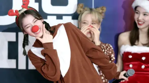 Sana from TWICE pictured at a photocall in Seoul in 2019. She is wearing a brown and white costume and reindeer antlers and a red nose. She is leaning to one side smiling at the camera. There are other people in Santa outfits in the background. 