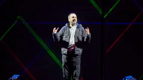 David Levene/Bradford City of Culture 2025 Steven Frayn stands on stage with his hands gesturing he is a man with short brown-grey hair. He is wearing black cargo trousers, a grey jumper and a black puffer jacket. Behind him are coloured lights.