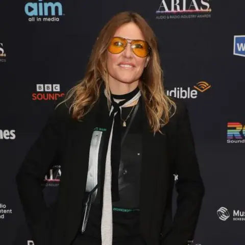 Getty Images Cerys Matthews wearing yellow sunglasses. She has long blonde hair and bits her lower lip as she smiles at the camera. She is wearing a black blazer, with a black and white tie.