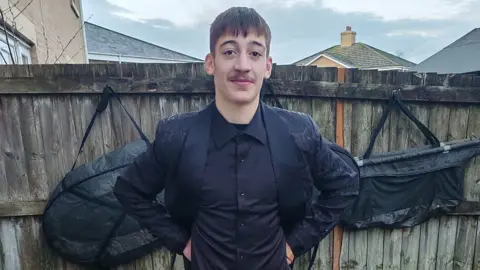 Devon and Cornwall Police A picture of Taylor wearing a black suit jacket and a black shirt while standing in front of a garden fence. He has a short moustache and short dark hair.