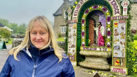 A woman with blonde long hair and a blue raincoat. Behind her is a large stone well surrounded by a huge floral display. The display includes an image of Jesus made from petals and natural materials. The words "I stand by the door and knock" are included in the design. On the columns of the design are images of Winnie the Pooh and other characters.