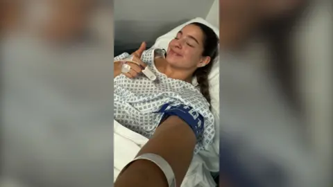 A woman with long brown hair in a plait is taking a selfie in a hospital bed. She has her eyes closed and is giving a thumbs up.