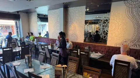 Shababs An Indian restaurant with customers sat waiting to eat their meals. A female employee in a purple uniform can be seen walking around. There are patterns on the wall and mirrors.