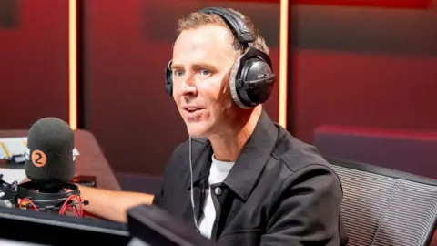 BBC Scott Mills in the Radio 2 studio wearing headphones and a black jacket