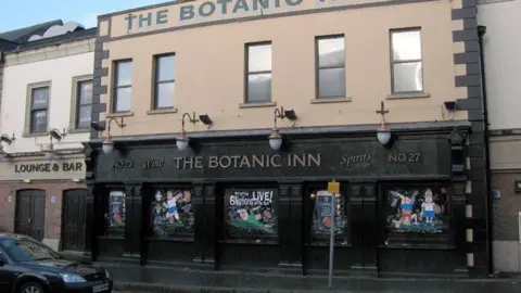 The Botanic Inn is a yellow and green building. It has black flags outside which read "Guiness". There are a number of hanging baskets on the corners of the building. The flowers are pink and yellow. 