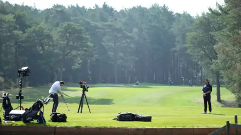 Getty Images A person hitting a golf ball down a long fairway with trees lining either side. He is facing away from the camera, and there are TV cameras filming him. A man is stood to the right watching.