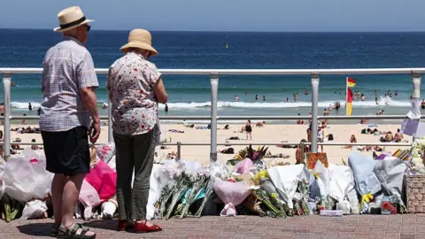 Two people stand in front of floral tributes on a paved path with a beach in the background