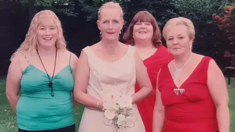 Family Photo Four women stood in a row at a wedding - Scott's mother is wearing the bridal dress and holding the bouquet.