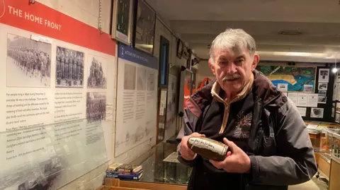 Colin Durward is a man in his 60s who is standing in a museum holding a second world war era bomb. He has grey hair and a moustache and is wearing a brown jacket.