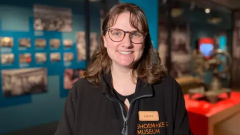 Laura is standing in the museum in front of different displays. She is wearing a black fleece, glasses and is smiling at the camera.