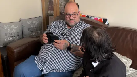 Christopher talking to Lilly on their sofa. Christopher is wearing a blue checked shirt and blue jeans, while holdig a mug and his phone. Lilly has her back to the camera.