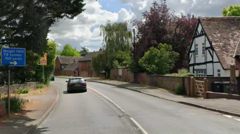 Google A curved road with trees on either side and a black and white timber framed building on the right and a car travelling on the left hand side of the road