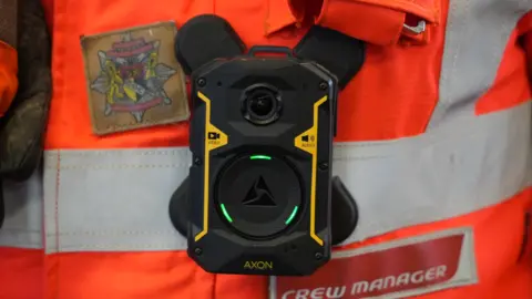 A close-up of a body-worn camera, positioned on an orange and reflective silver top. 