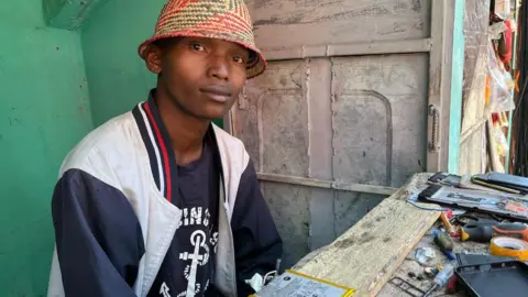 Sami Awami/BBC Laza Brenda, a young man wearing a straw hat, sits at his mobile phone repair booth.