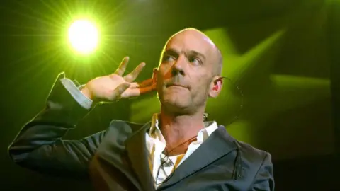 PA Media REM frontman Michael Stipe performs on stage. He has one hand raised to his ear as he listens to the crowd. He holds a microphone stand in the other hand. He wears a suit jacket with a white shirt underneath.