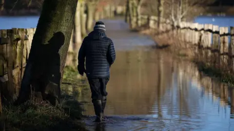 A man wearing gumboots walks along a flooded walkway between wooden fences