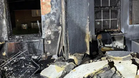 A burnt out room with bits of blackened debris. The wall can be seen split into pieces and scattered across the ground. The windows are also blown out. 