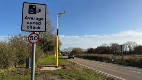 A speed limit sign bearing the numeral 50, in a red circle. Above that is a rectangular sign, showing a pictogram of a camera, with the text "Average speed check" within, and behind that, a yellow pole which arcs to the right at the top, upon which is a camera. Traffic can be seen on a single-carriageway road. Hedges can be seen either side of the road.