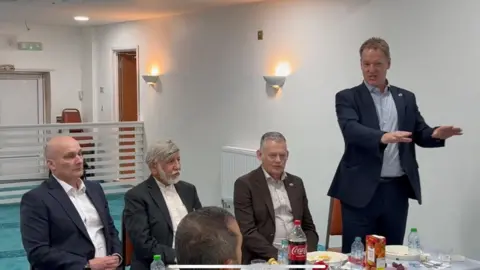 Abdul Choudhuri Three smartly dressed men sit at a table with food on it in a mosque. A fourth man, on the right, is standing up making a speech.