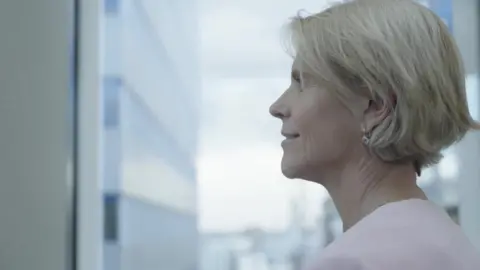 Meagen Burnett, a woman with short blonde hair wearing a light pink top, looking out of a window in a high rise office building