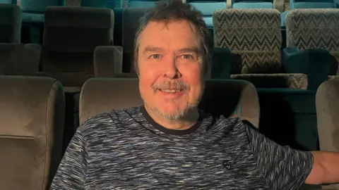 A man with short dark hair, a greying beard and moustache, is sitting in a cinema seat wearing a blue, black and white patterned t-shirt