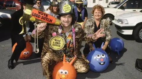 BBC/ Willie Smith Screaming Lord Sutch of the Monster Raving Loony Party, at their 1990 party conference. He wears a leapoard skin suit with a matching leopardskin top hat and is sitting astride an orange spacehopper. He is wearing several giant yellow and black rosettes and carries a golden loudhailer, all with the slogan Vote Lord Sutch. He is surrounded by a group of supporters also riding spacehoppers in a car park.