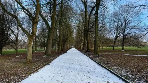 Snow has settled on a path lined by trees in Longford Park, Stretford 