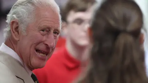 Close up of King Charles smiling wearing a beige suit during a visit to Morecambe Fire Station in 2022 and talking to a girl with long brown hair captured from behind.