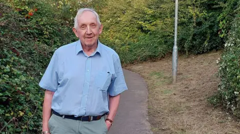 Andrew Gravells Andrew Gravells wearing light green chinos, a dark belt and a short-sleeved blue shirt. He has short white hair and is smiling at the camera, with his left arm in his trouser pocket. He is standing on a curved footpath beside overgrown bushes. 