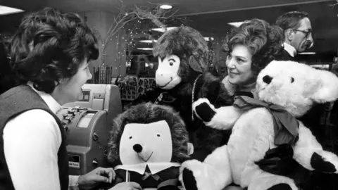Getty Images A woman with brown curly hair buys toys for her grandchildren during a shopping spree Rackhams Department Store. Birmingham, West Midlands, November 1976. 