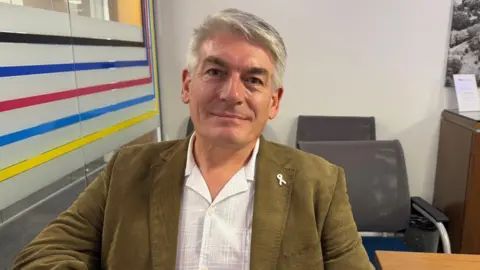 Simon Nash is pictured wearing a green blazer and a white shirt. He is sat in a bright office setting.