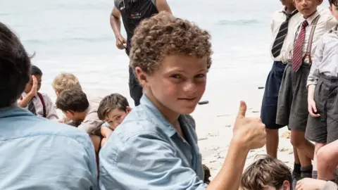 BBC/Eleven/J Redza A boy sitting on a beach, surrounded by peers, holding his thumb up and smiling sat the camera.