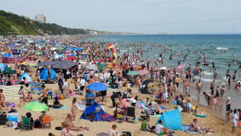 A beach is filled with thousands of sunbathers, parasols and tents as crowds flock to the beach in the sunshine. Many are paddling and swimming in the sea. red and Yellow lifeguards flags can be seen standing in the sand.
