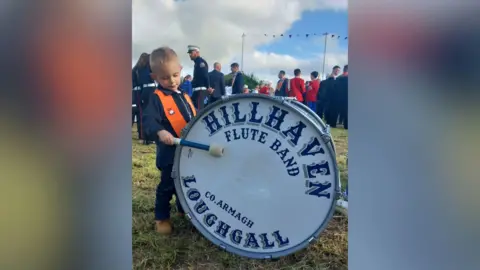 Claire Reaney A small blonde boy wearing a navy tracksuit and orange sash bangs a lambeg drum which says 'hill haven flute band county Armagh loughgall'