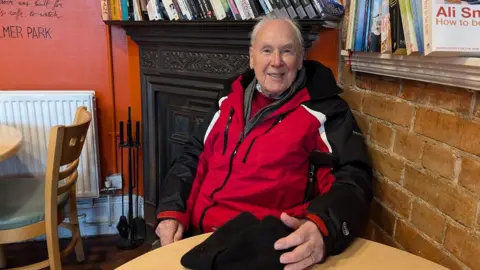 A man with white hair and wearing a red anorak sitting at a table in a cafe. There's a shelf of books to his right, a black fireplace behind him and a red wall to the left. He's smiling at the camera.