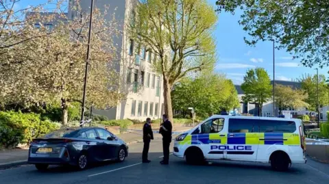 Police tape closes off the road. Two officers can be seen stood next to a police vehicle. The area is leafy with a pale building in the background.