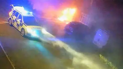 Flames engulf the front of a house with a police car parked outside.