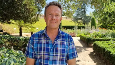 Bishop's Palace Wearing a short sleeved blue, checked shirt, James smiles at the camera. He stands on a path within a well kept garden, with a small hedge lining the path on his left and other plants on his left. Several trees also surround him.