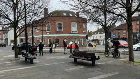 George Carden/BBC A shot of a town square in Chichester with bences and trees around a paved aread with a large municipal building in the background 